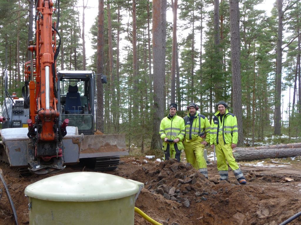 Till vänster i bilden syns en grävmaskin. Till höger står tre män i signalgula arbetskläder. I förgrunden syns överdelen av en pumpstation. Bakom grävmaskinen och männen växer tallar.