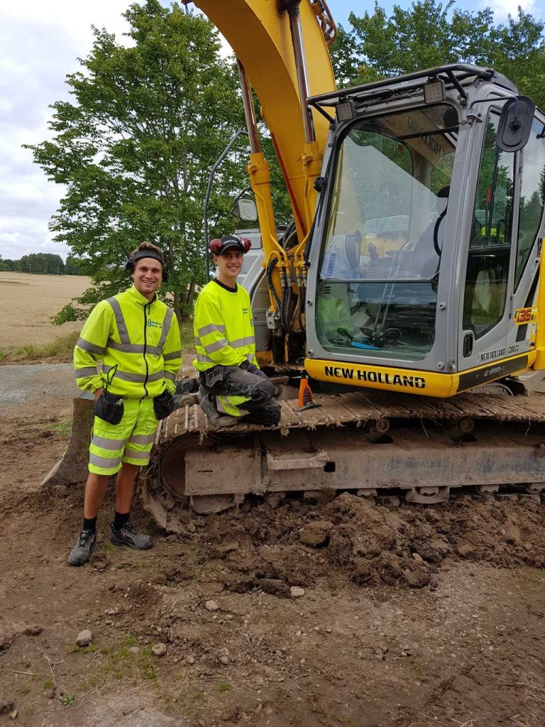 Två glada män från Götene Grävtjänst vid en grävmaskin av märket New Holland.